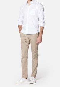 Bílá košile na knoflíky s náprsní kapsou, v kombinaci s béžovými chinos kalhotami a bílými teniskami. Materiál vypadá hladce a má klasický střih.