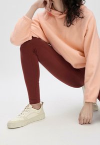 Felpa con cappuccio in pile rosa chiaro con colletto alto, abbinata a leggings color ruggine e sneaker color crema. Superficie piatta, sfondo minimale.