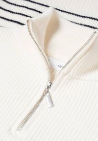 Maglione bianco a costine con colletto a righe nere, dotato di una cerniera argentata e di un'etichetta; texture morbida e design informale.