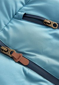 Ein strukturierter blauer Stoff mit zwei Reißverschlusstaschen, die mit bronze-farbenen Beschlägen und braunen Leder-Zip-Pulls versehen sind. Glatte, glänzende Oberfläche.