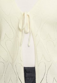 Cardigan de couleur crème avec des découpes en forme de cœur et une fermeture à nouer à l'avant. Texture en tricot douce avec un col rond.