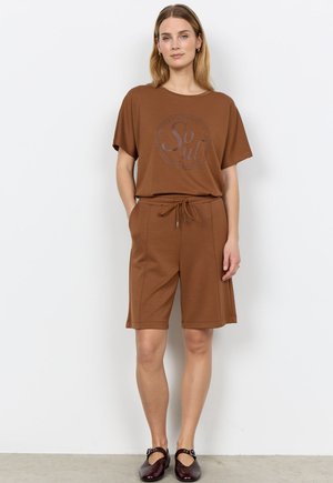 Eine Frau steht vor einer weißen Wand und trägt ein braunes, lockeres T-Shirt mit dem Aufdruck „So Sul“, passende Shorts mit Kordelzug und dunkelbraune Mary-Jane-Schuhe.