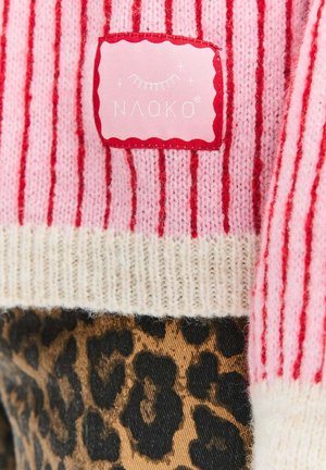 Růžový pruhovaný svetr s bílým žebrovaným lemem a červenou štítkem s nápisem "NAOKO." Dolní část zdobí leopardí vzor.