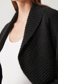 Černý pletený kardigan s vroubkovanou texturou a šálovým límcem, vrstvený přes bílý top, zaměřený na design a materiál oděvu.