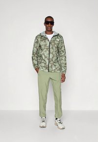 Lagana, zelena maskirna jakna s kapuljačom, patentnim zatvaračem i odgovarajuće svijetlozelene hlače. Model nosi crne sunčane naočale i sive tenisice.