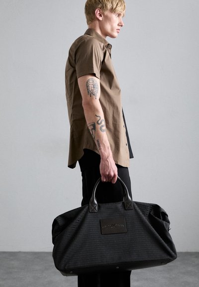 Sac de sport noir avec un tissu texturé, des accents en cuir et un patch logo. Dispose de poignées robustes et d'une forme spacieuse.
