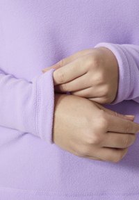 Polarowy półgolf w jasnym lawendowym kolorze o miękkiej teksturze. Zbliżenie na mankiety z kontrastowym zszyciem oraz dłonie dostosowujące rękaw.