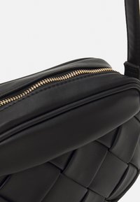Sort, vævet læder crossbody taske med guld lynlås og polstret detaljering synlig på toppen og fronten.