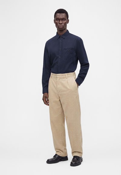 Man wearing a dark blue buttoned shirt, beige corduroy pants, and black shoes standing with one hand in pocket against a white background.