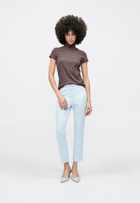 Sieviete ar cirtainiem matiem, valkājot rakstainu īsroku kreklu, gaiši zilas, piegrieztas bikses un pelēkus smailus papēžus, stāv pret baltu fonu.