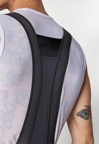 Schwarzes Mesh-Tanktop mit einer strukturierten weißen Unterlage. Verfügt über verstellbare Träger, einen glatten Ausschnitt und ein sichtbares Tattoo am Arm.