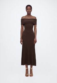 Γυναίκα στέκεται όρθια φορώντας καφέ midi φόρεμα με ριμπ ύφανση και ακάλυπτους ώμους, καθώς και χρυσά ψηλοτάκουνα σανδάλια, μπροστά από ένα απλό λευκό φόντο.