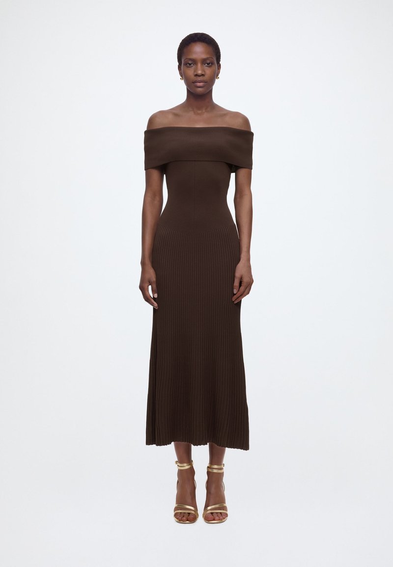 Γυναίκα στέκεται όρθια φορώντας καφέ midi φόρεμα με ριμπ ύφανση και ακάλυπτους ώμους, καθώς και χρυσά ψηλοτάκουνα σανδάλια, μπροστά από ένα απλό λευκό φόντο.