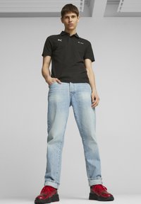Czarna koszulka polo z kołnierzykiem, wąskie niebieskie dżinsy i czerwone sneakersy w wzory. Koszulka ma logo PUMA oraz branding Mercedes-Benz.