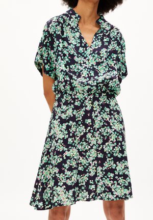 Frau trägt ein kurzärmeliges schwarzes Kleid mit grün-weißem Blumenmuster und leicht ausgestelltem Rock.