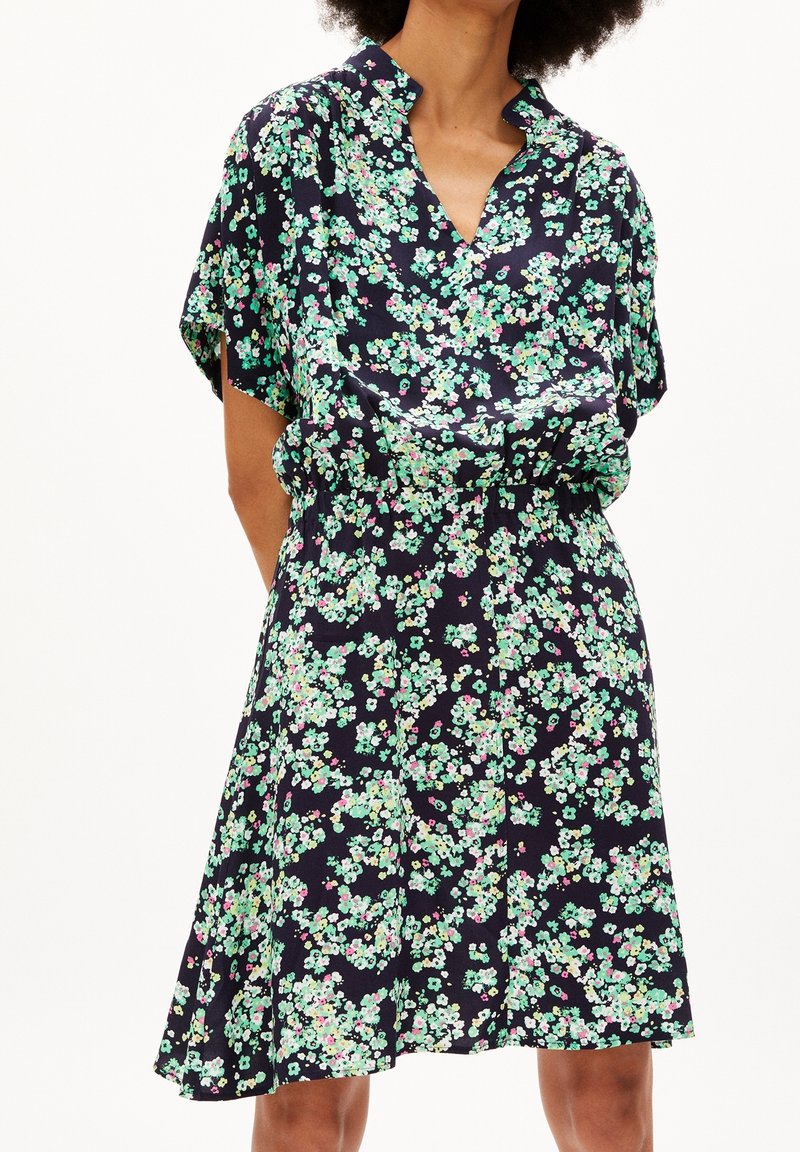 Femme portant une robe noire à manches courtes avec un motif floral vert et blanc et une jupe légèrement évasée.