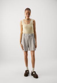 Knit tank top vaale yellow rang mein scalloped edges ke saath, halka gray tailored shorts aur bhuri jootiyon ke saath, jo moti sole ke saath hai.