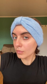 Lyseblå strikket hodebånd med vridd design, båret på en persons hode, komplementert med en svart skjorte og nøytral bakgrunn.