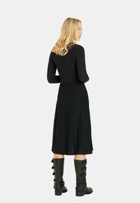 Schwarzes, plissiertes Kleid mit langen Ärmeln und strukturiertem Oberteil, kombiniert mit knielangen schwarzen Stiefeln mit dekorativen Hardware-Details.