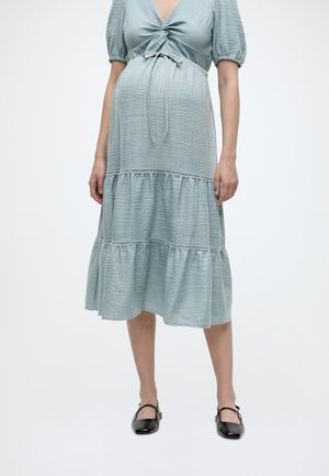 Femme enceinte portant une robe midi texturée bleu clair avec des manches bouffantes et des chaussures Mary Jane noires, debout devant un fond uni.