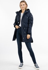 Marineblå parka med hætte, frontlynlås, justerbare snoretræk, to sidelommer og hvide detaljer. Båret over en hvid skjorte og mørke jeans.
