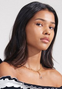 Guldkæde med vedhæng i trekantet form og geometrisk design. Modellen har mørkt, bølget hår og bærer en off-shoulder top.