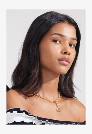 Zlatý řetízkový náhrdelník s trojúhelníkovým přívěskem s geometrickým vzorem. Modelka má tmavé, vlnité vlasy a nosí top s odhalenými rameny.