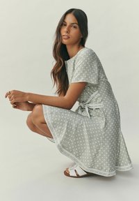 Hellgraues Kleid mit weißen Polka-Dots, kurzen Ärmeln und tailliertem Gürtel. Es hat einen ausgestellten Saum und dekorative Verzierungen. Wird mit weißen Sandalen getragen.