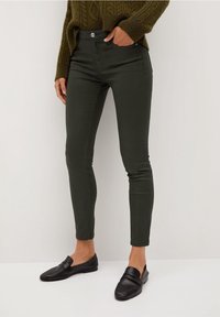 Mujer vestida con pantalones oscuros verde ajustados, suéter de punto en color verde oliva y mocasines negros, de pie contra un fondo blanco.