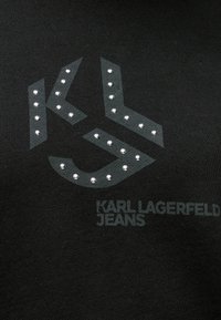 Fekete textil, domináló szürke logóval, mely rhinestone-okkal díszített. A logo alatt diszkrét tervezésben olvasható a "KARL LAGERFELD JEANS" felirat.