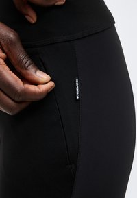 Χέρι που κρατάει και ανοίγει μια κάθετη τσέπη με φερμουάρ σε μαύρο εφαρμοστό αθλητικό κολάν με μικρή ετικέτα λογότυπου.