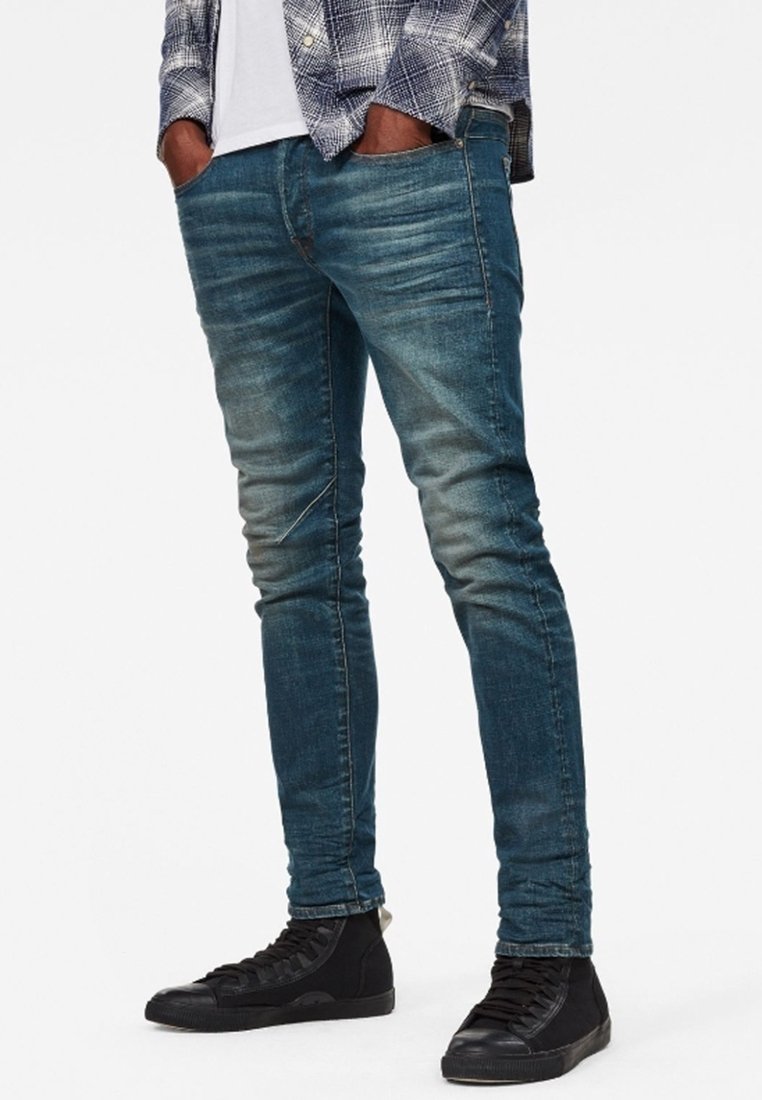 Jeans skinny de mezclilla azul con un acabado desgastado, que presentan un diseño clásico de cinco bolsillos y una textura ligeramente rasgada. Combinados con zapatillas negras.