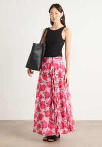 Haut sans manches noir associé à une jupe maxi fleurie rose avec des volants et de grands motifs de roses rouges, complétée par un sac fourre-tout noir.