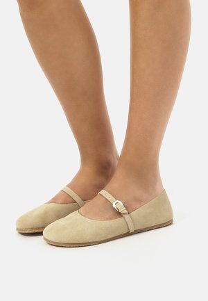 Le ballerine in suede beige presentano una punta tonda, una fascia alla Mary Jane con fibbia e una suola in gomma naturale testurizzata.