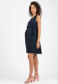 Vestito premaman plissettato blu navy con vita annodata e design senza maniche; abbinato a tacchi rosa con cinturini. Tessuto morbido con una silhouette fluida.