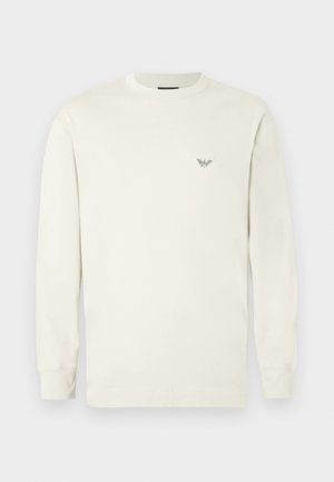 Svijetlo bež majica dugih rukava od pamuka. Ima okrugli izrez i mali izvezeni logo na prsima. Glatka tekstura.