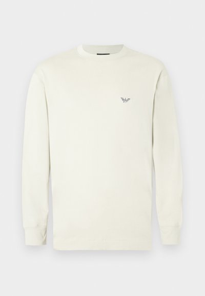 Gaiši bēša kokvilnas garroku krekls. Tai ir apaļš kakla izgriezums un neliels izšūts logo krūtīs. Gluda tekstūra.