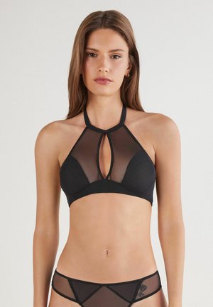 Vrouw met lang bruin haar in een zwarte, doorschijnende halterbralette en bijpassende doorschijnende slip, staand tegen een effen achtergrond.