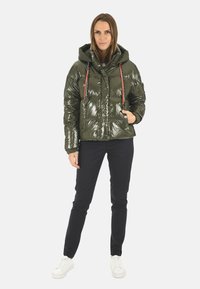 Veste matelassée verte avec une finition brillante, capuche et cordons de serrage rouges, associée à un pantalon noir et des baskets blanches. Comprend des poches zippées.