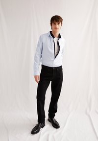 Lichtblauw button-up overhemd over een wit T-shirt, gecombineerd met zwarte jeans en zwarte schoenen. Het overhemd heeft een contrasterende donkere kraag.