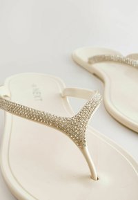 Sandales plates blanches avec des brides en forme de Y ornées de strass sur un fond blanc uni.