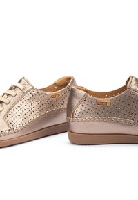 Zapatos de cuero metálico dorado con diseño perforado, cordones en la parte delantera y suelas de goma en contraste en color marrón. Presenta branding en los laterales y detalles de costura.