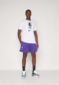Valge lühikeste varrukatega T-särk musta NBA logo ja Nike swooshiga; lillad võrkpüksid NBA logoga; mustad ja valged sportlikud jalanõud; mustad sokid.