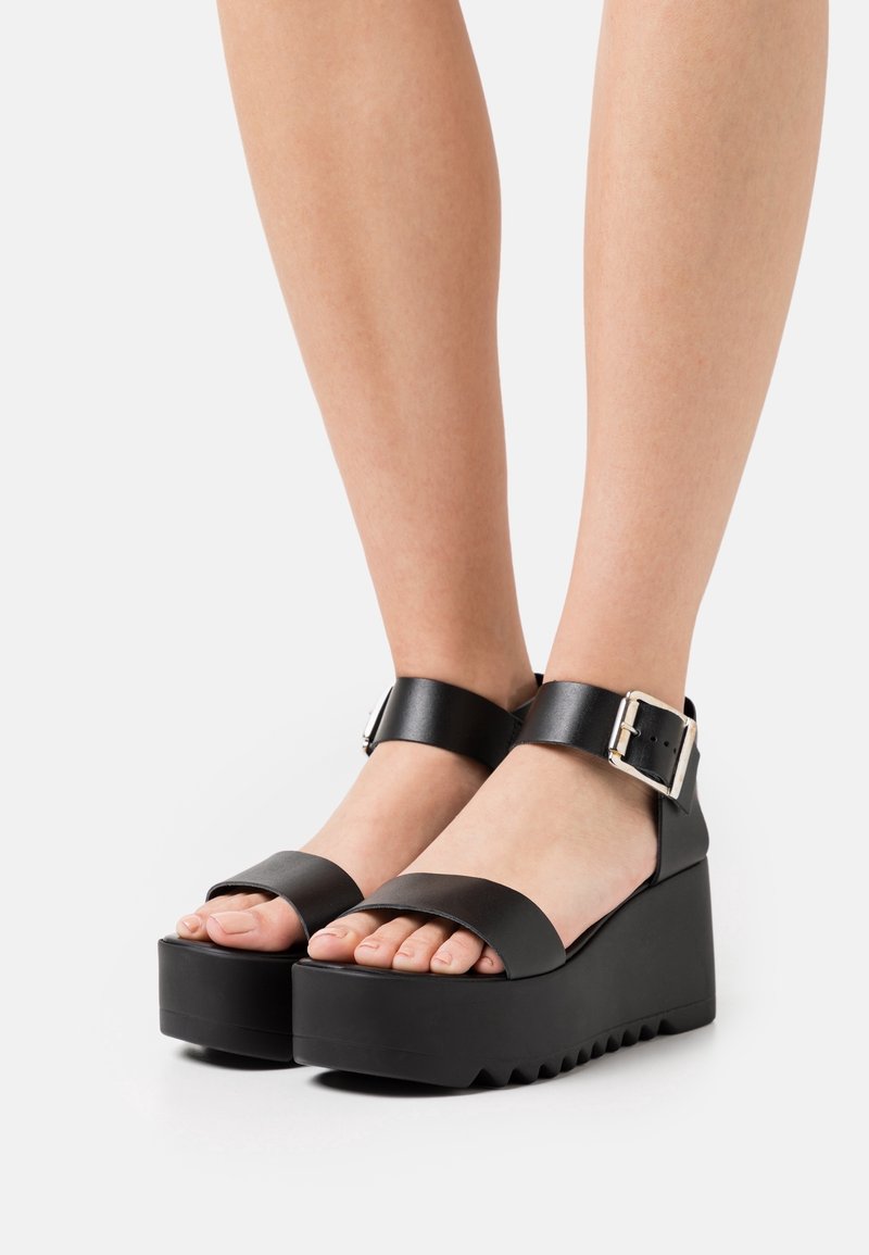 Sandalias negras de plataforma con correas anchas, detalle de hebilla y suelas gruesas. Material de cuero suave con una base texturizada.