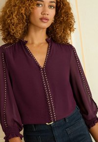 V-veida blūze bagātīgā purpursarkanā krāsā ar volānu apkakli, caurspīdīgām bumbierveida piedurknēm un dekoratīviem zeltainiem kniedēm gar plātni un piedurknēm.