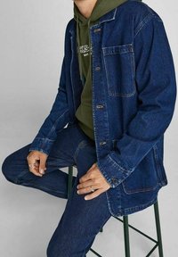 Giubbotto di jeans blu scuro con chiusura a bottoni e tasche frontali, indossato sopra un hoodie verde. Modello seduto su uno sgabello.