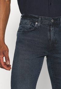 Vaqueros de mezclilla azul oscuro con un corte slim, que presentan un diseño clásico de cinco bolsillos, cierre de botón en la parte frontal y un ligero desgaste en la superficie de la tela.