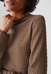 Donna con capelli ricci che indossa un top lungo marrone con texture, maniche a sbuffo e colletto, con una mano appoggiata vicino alla clavicola.