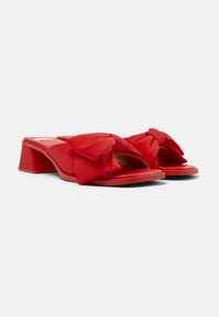 Red heeled mules made of satin, featuring a large bow at the front, open toe design, and a square block heel. Smooth texture throughout.