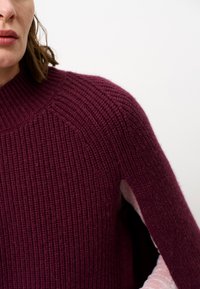 Bordeaux ribbelgebreide trui met een hoge kraag en raglanmouwen, met een textuurafwerking. Achtergrond is neutraal.